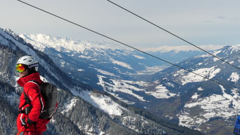 Mitteltotale-Aufnahme-Von-Seilbahnen,-Die-An-Einem-Strahlend-Sonnigen-Tag-In-Wagrain-Bergauf-Und-Bergab-Fahren,-Mit-Einem-Mann-In-Skiausrüstung-Im-Vordergrund-Und-Wunderschönen-Schneebedeckten-Bergen-Im-Hintergrund