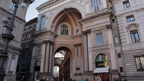 Galleria-Vittorio-Emanuele-II-entrance-from-the-Piazza-della-Scala-in-Milan-Italy,-wide-dolly-in-tilt-up-shot-during-daytime-with-people-walking