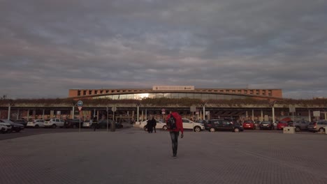 Man-with-red-coat-and-backpack-walks-toward-Train-Station-in-Seville,-Spain