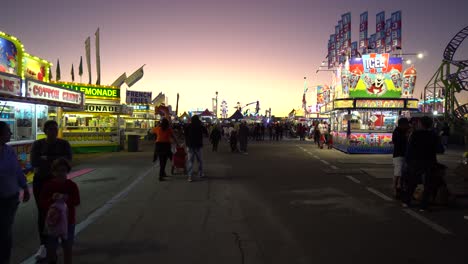 Toma-De-Seguimiento-De-Personas-Caminando-En-La-Feria-Estatal-De-Florida-Con-Puestos-De-Comida-Y-Atracciones-A-La-Vista-Durante-El-Crepúsculo.