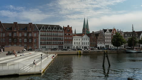 Gente-Disfrutando-Del-Sol-En-El-Muelle-Del-Río-Trave-Con-Las-Arquitecturas-Del-Casco-Antiguo-Al-Fondo-En-Lübeck