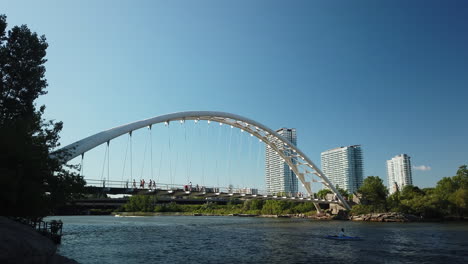 Wide-shot-of-people-crossing-the-HumBer-Bay-Arch-Bridge,-with-lone-kayaker-underneath