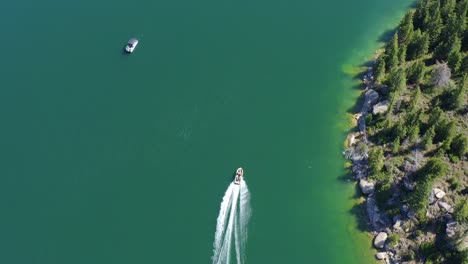 Aerial-bird's-eye-view-of-a-boat-motoring-along-the-shoreline-of-a-clear,-blue-lake