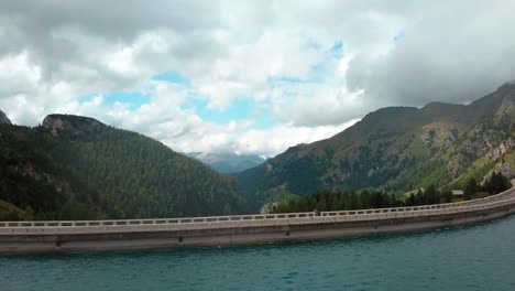 Fedaia-Dam-lake-in-the-Dolomite-mountain-area-of-northern-Italy-with-a-person-admiring-view-from-the-railing-structure-to-the-right,-Aerial-Drone-flyover-view