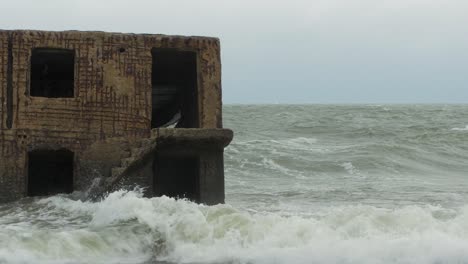 Big-stormy-waves-breaking-against-abandoned-seaside-fortification-building-ruins-at-Karosta-Northern-Forts-in-Liepaja,-Baltic-sea-coastline,-wave-splash,-overcast-day,-medium-shot