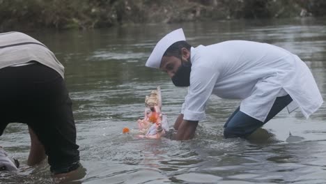 Indischer-Mann-Mit-Maske-Und-Sozialem-Distanzierungs-Coronavirus,-Der-In-Einem-Fluss-Steht-Und-Ganpati-Ganesh-Idol-In-Zeitlupe-Eintaucht