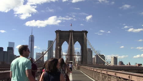 Los-Turistas-Cruzan-El-Puente-De-Brooklyn-En-Un-Día-Soleado,-La-Torre-De-La-Libertad-De-Manhattan-En-Segundo-Plano