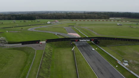 Coches-Acelerando-A-Lo-Largo-De-La-Pista-De-Carreras-De-Snetterton-Bajo-El-Puente-Con-El-Cartel-De-Snetterton