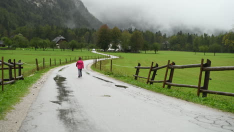 Little-girl-walking-in-rain-on-country-road,-farmhouse-in-background,-alpine-valley,-4k-steady-shot,-from-behind-facing-away