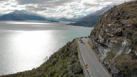 Motorhome-driving-in-the-mountains-along-the-coast-of-Lake-Wakatipu,-Queenstown,-New-Zealand-with-mountains-fresh-snow-in-background---Aerial-Drone