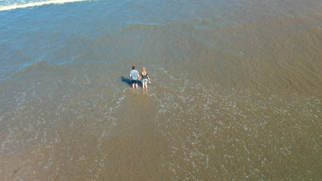 Aerial-revealing-shot-of-a-couple-standing-hand-in-hand-in-the-ocean-at-the-ninety-mile-beach-with-waves-rolling-over-the-beach,-New-Zealand