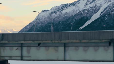 Bridge-of-frozen-river,-aerial-rise-revealing-cars-traveling-road-and-sun-rising-on-Alaskan-mountains,-Anchorage