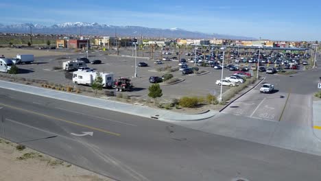 Luftaufnahmen-Eines-Walmart-Parkplatzes-In-Adelanto,-Kalifornien,-Mit-Geparkten-Freizeitfahrzeugen,-Fahrenden-Autos,-Gehenden-Menschen,-Geschäften-Und-Schneebedeckten-Bergen-Von-Angeles-Im-Hintergrund
