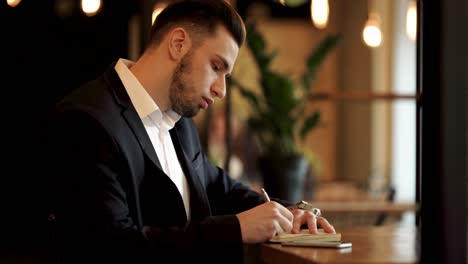 Stout-handsome-man-with-black-beard,-whiskers-in-classic-black-suit-and-white-shirt-sitting-in-a-cozy-cafe-flipping-pages-of-notebook,-holding-pen,-starting-talking-on-phone