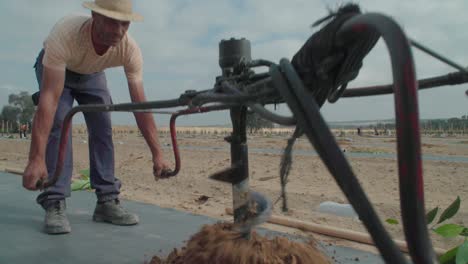 Earth-auger-drilling-sapling-hole-in-plastic-mulch,-Slow-Motion