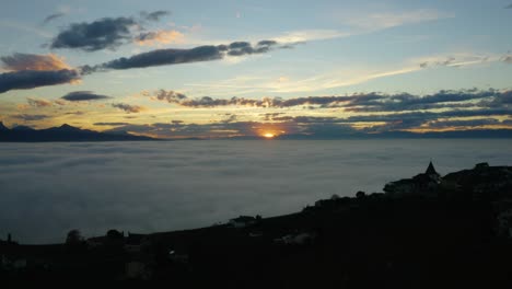 Aerial-shot-passing-small-village-at-the-edge-of-a-sea-of-fog-covering-Lake-Léman,-looking-like-the-end-of-a-magical-world-at-sunset