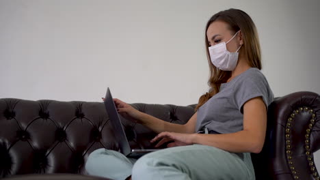 Telearbeit-Während-Der-Coronavirus-Pandemie.-Frau,-Die-Von-Zu-Hause-Aus-Mit-Dem-Laptop-Arbeitet,-Eine-Medizinische-Gesichtsmaske-Trägt-Und-Auf-Einem-Sofa-Sitzt.