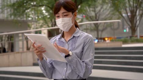 Japanese-business-woman-reading-a-contract-document-wearing-a-medical-face-mask-outdoors.-New-employee-hiring-during-the-covid-19-pandemic.