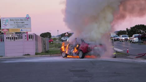 Feuerwehrmann-Löscht-Brennendes-Auto-Auf-Parkplatz-Mit-Wasserschlauch,-Massive-Flammen-–-Rauchwolken