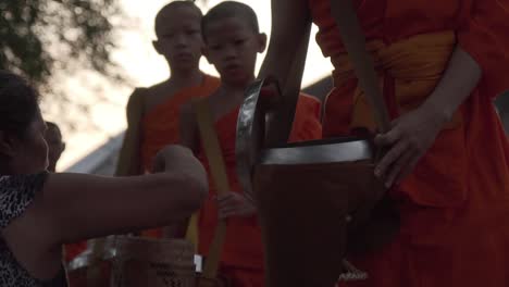 Junge-Buddhistische-Mönche-In-Traditionellen-Orangefarbenen-Gewändern-Almen-Morgens-Auf-Der-Straße-Und-Eine-Asiatische-Laotische-Frau-Füttert-Mönche-Mit-Reis-In-Einem-Bambuskorb