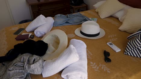 Handheld-Shot-of-a-Messy-Hotel-Room-Bed-With-Sun-Hats,-Sun-Glasses,-Passports,-Mobile-Phone,-Towels,-Jeans-Skirt,-Controller,-Keys,-Pillows-and-a-Wooden-Drawer-in-The-Background