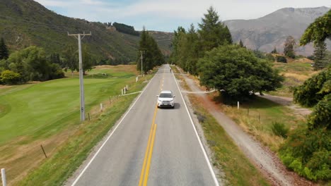 SLOWMO---Luxury-suv-car-driving-along-golf-course-in-Arrowtown,-New-Zealand-while-golfers-crossing-road---Aerial