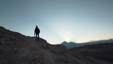 Hiker-silhouette-walking-down-a-mountain-on-desert-at-sunrise-with-volcanoes-in-the-background