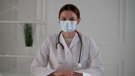 Young-pretty-female-doctor-with-a-face-mask-looks-at-camera-and-smiles-at-the-office