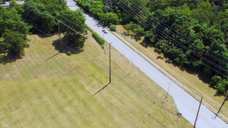 bird's-eye-view-over-power-lines,-a-green-field---trees,-as-a-white-car-drives-by-below