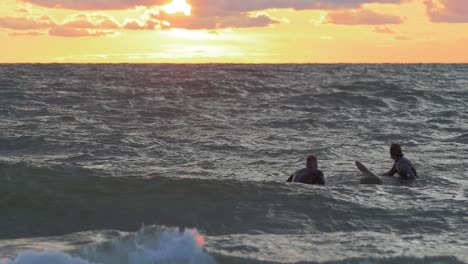 Surfers-on-surfboards-paddling-over-waves-near-the-Baltic-sea-Karosta-beach-at-Liepaja-during-a-beautiful-vibrant-sunset-at-golden-hour,-medium-slow-motion-shot-from-a-distance