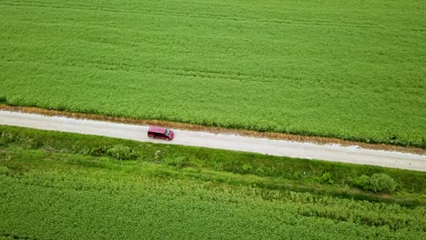 Aerial-landscape-sideway-shot-of-claret-van-driving-on-gravel-road-between-green-agricultural-fields-hungary-europe