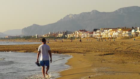 Young-man-walking-barefoot-on-shoreline-of-sandy-beach,-coastal-town,-Spain
