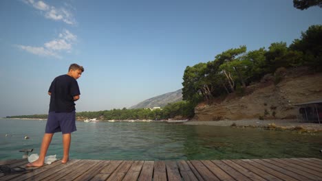 Barefoot-young-man-stands-on-end-of-wooden-dock-with-back-to-camera