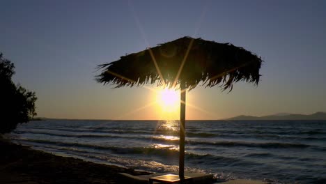 Ein-Sonnenschirm-Aus-Palmblättern-Und-Sonnenliegen-Im-Sonnenuntergang-Am-Strand-Von-Kos-An-Der-Ägäis