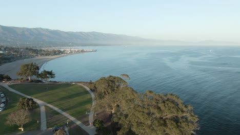 High-altitude-aerial-over-coastline-city-park-during-sunset-with-trees-and-the-Pacific-ocean-with-mountains-in-the-background,-in-Santa-Barbara,-California,-USA