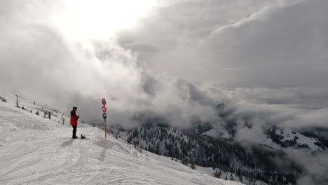 Weitwinkelaufnahme-Eines-Mannes,-Der-Neben-Einem-Schild-Steht-Und-Skiausrüstung-Trägt,-In-Einem-Skigebiet-In-Wagrain-Mit-Wunderschönen-Schneebedeckten-Bergen-Und-Dramatischen-Wolken-Im-Hintergrund