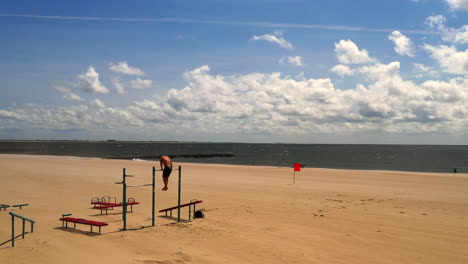 drone-camera-steady-shot,-as-a-young-man-exercises-on-a-pull-up-bar-on-Coney-Island-beach-in-Brooklyn,-NY-at-a-workout-station-with-the-sandy-beach,-the-Atlantic-Ocean,-the-blue-sky---horizon-in-view