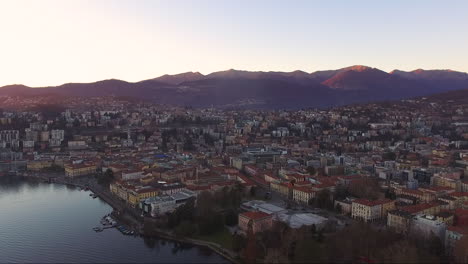 A-drone-view-of-a-beautiful-city-surrounded-by-mountains,-next-to-a-lake,-during-a-sunset-in-autumn