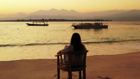 Melancholic-girl-sitting-on-chair-over-sandy-beach,-watching-silhouette-of-boats-floating-on-calm-lagoon,-Indonesia