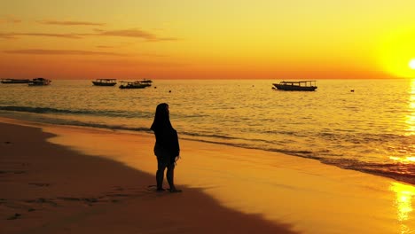 Young-woman-enjoying-beautiful-seascape-with-orange-sky-and-yellow-sun-reflecting-on-calm-bay-with-silhouettes-of-boats,-Bali