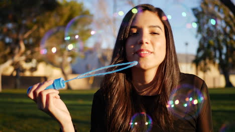 Attractive-hispanic-woman-blowing-dreamy-and-colorful-bubbles-while-smiling-and-looking-playful-outdoors-at-sunset