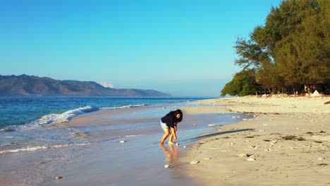 Lonely-girl-playing-barefoot-with-waves-of-blue-sea-washing-quiet-sandy-beach-of-tropical-island,-clear-bright-sky-in-Philippines