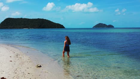 Young-woman-in-short-jeans-washing-feet-on-shallow-calm-lagoon-with-clear-water-on-shore-of-tropical-island-with-white-sandy-beach-in-Thailand
