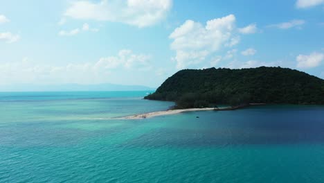 Paradise-tropical-island-with-hill-covered-in-lush-vegetation,-white-sandy-beach-surrounded-by-blue-turquoise-sea-water-under-bright-sky-in-Thailand