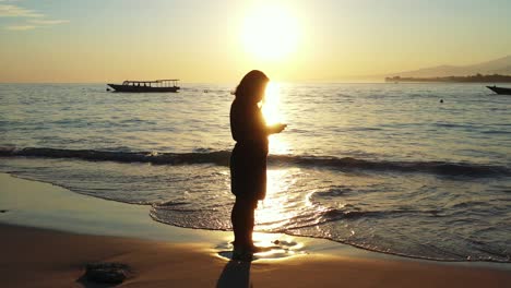 Silhouette-Eines-Mädchens,-Das-Bei-Sonnenuntergang-Mit-Dem-Smartphone-Meereswellen-Fotografiert,-Während-Sich-Das-Wunderschöne-Sonnenlicht-Auf-Der-Lagunenoberfläche-Spiegelt