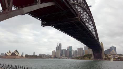 Underside-view-of-Harbour-Bridge-with-Opera-House-and-downtown-buildings-in-the-distance-on-an-overcast-day,-Pan-right-shot