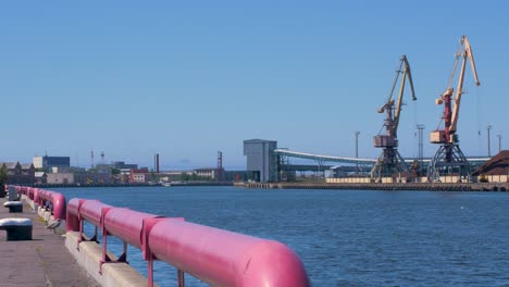 Port-cranes-and-harbor-warehouses-in-sunny-calm-summer-day-at-Port-of-Ventspils,-Venta-river-bank,-wide-shot-from-a-distance