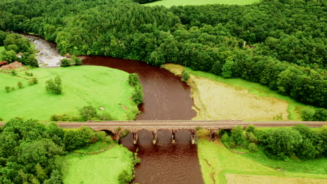 Aerial-wide-shot-of-a-railway-viaduct-crossing-a-river-surrounded-by-green-countryside-and-forest,-bright-daylight
