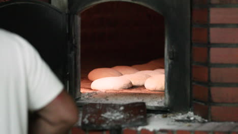A-Male-Baker-In-White-Shirt-Placing-The-Bread-Dough-Inside-A-Traditional-Stone-Oven---Making-Bread-In-Bakery---Medium-Shot