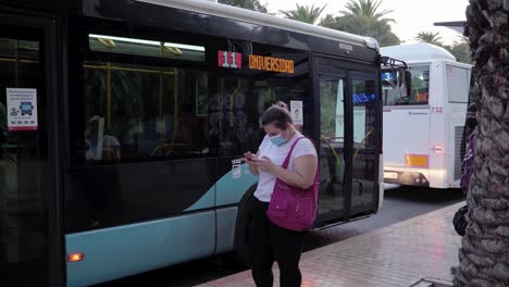 Viajero-Femenino-Con-Máscara-Usando-Un-Teléfono-Inteligente-Mientras-Está-De-Pie-En-Una-Parada-De-Autobús-En-Málaga,-España---Autobuses-Que-Salen-Durante-El-Día---Tiro-Lateral